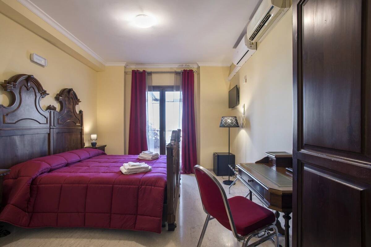 A hotel room with a bed, a desk, a chair, an air conditioning unit, and a window with curtains.