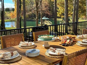 Una mesa de comedor de madera al aire libre con sillas, platos, tazones y vista a un lago y árboles.