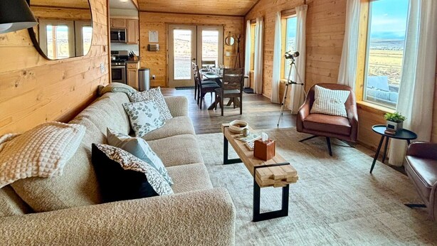 A cozy cabin interior with a wooden ceiling, a sofa, a dining table, and a view of the outdoors. - Explore Oregon Outback - Sauna - Hot Tub - Stargaze - WIFI - Bird Watching (Summer Lake)