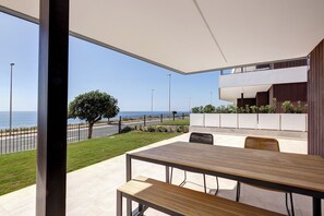 Eine moderne Terrassengruppe im Freien mit Holztisch und -stühlen, Blick auf das Meer, sowie ein Gebäude mit weißer Fassade und dunklen Holzelementen.