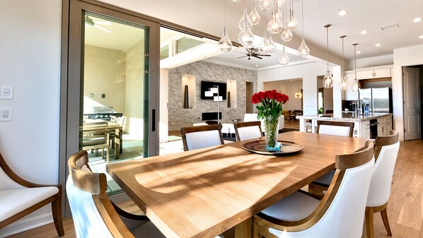 A modern dining area with a large wooden table, white upholstered chairs, and a vase of red flowers.