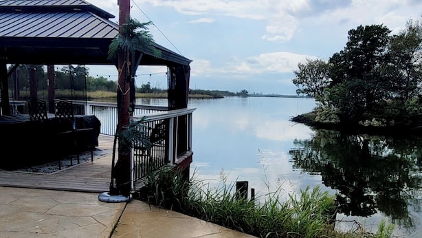 Un gazebo con tetto in metallo affacciato su un'acqua tranquilla, un vialetto in pietra e un pontile in legno con mobili da esterno.