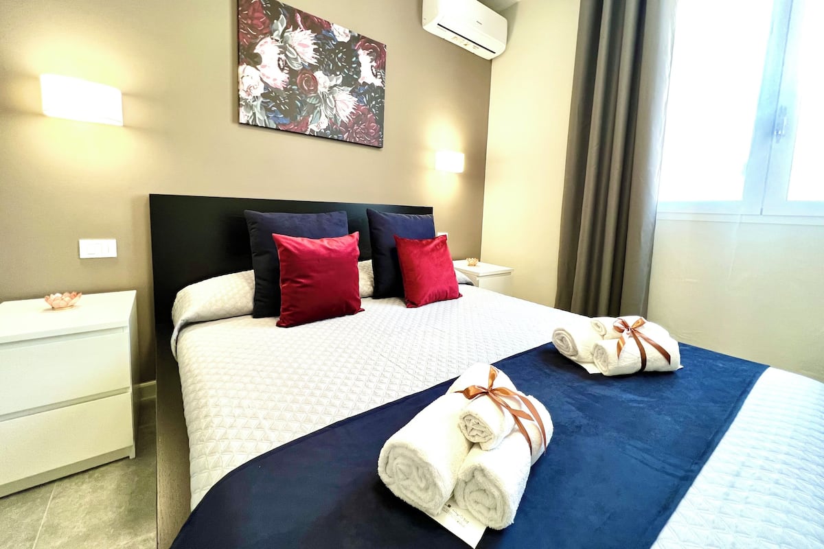 Habitación de hotel con cama, almohadas, mesita de noche, aire acondicionado de pared y ventana con cortinas.