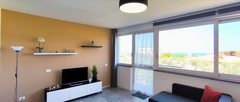 A modern living room with a TV, sofa, and large windows offering a view of the outdoors.