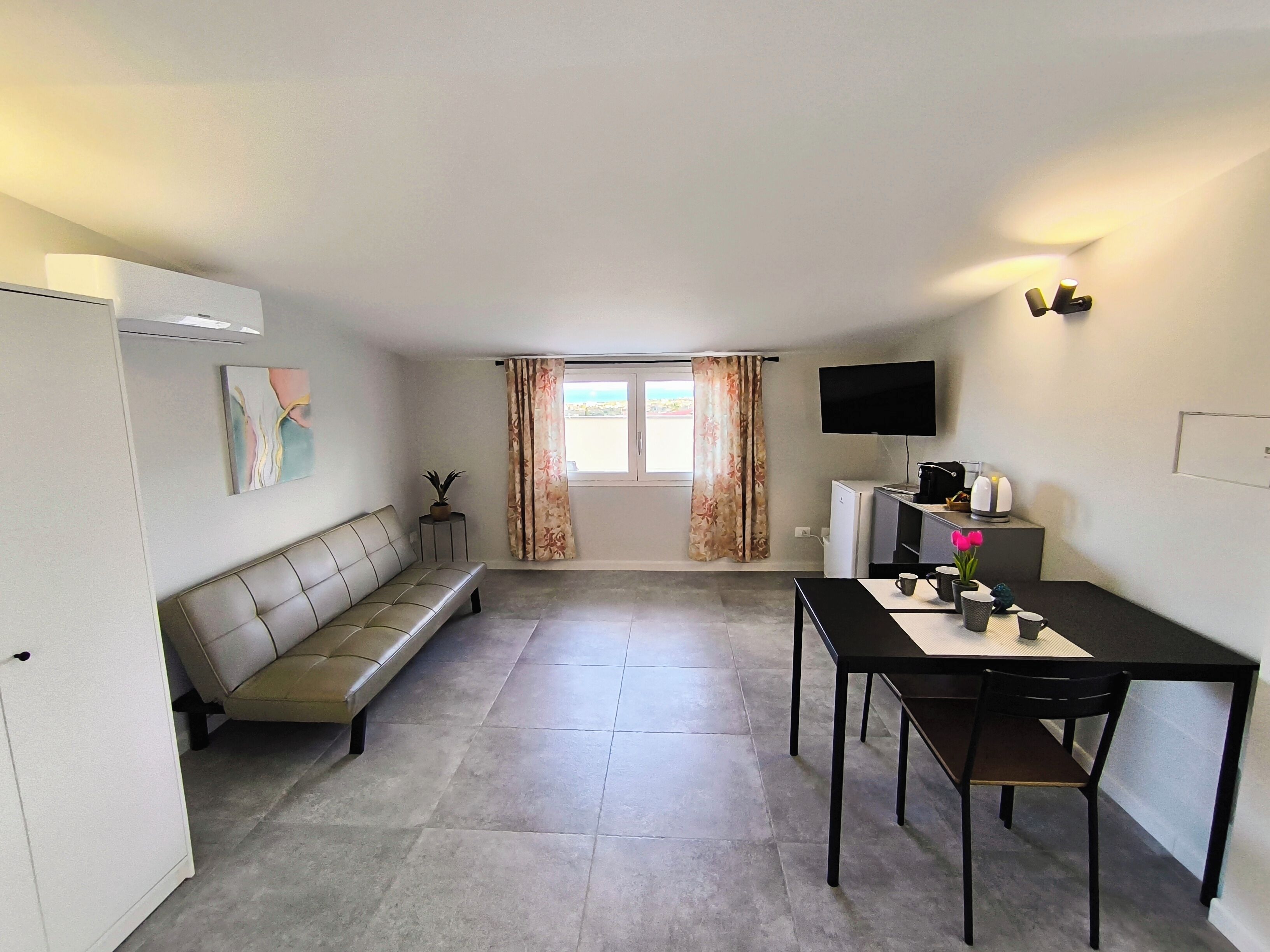 Una habitación pequeña y moderna con sofá, una mesa de comedor con sillas, televisión y ventana con cortinas.
