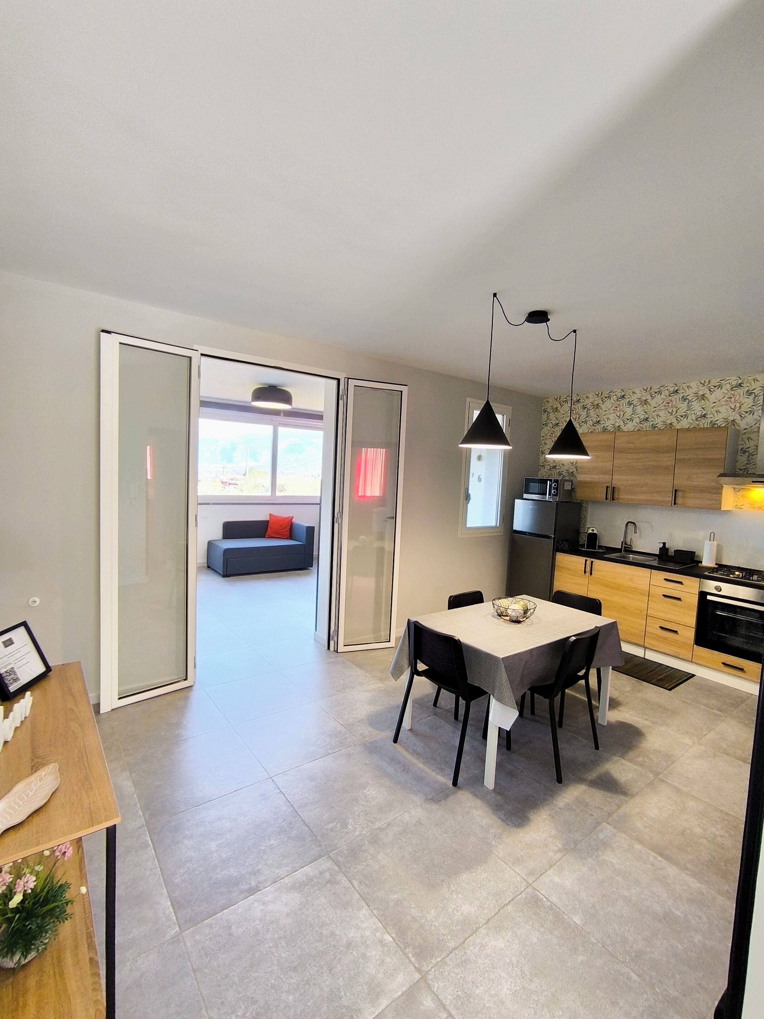 A modern kitchen with a dining area, a view through a glass door, and a small seating area with a red cushion.