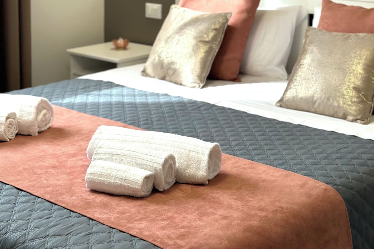 Una cama bien hecha con colcha azul, manta rosa y toallas blancas enrolladas.
