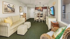 A compact living space with a sofa, ottomans, a dining area with chairs, a ceiling fan, and a TV.