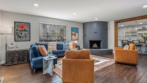 A modern living room with a fireplace, a blue sofa, an orange armchair, and a dining area visible through an open doorway.