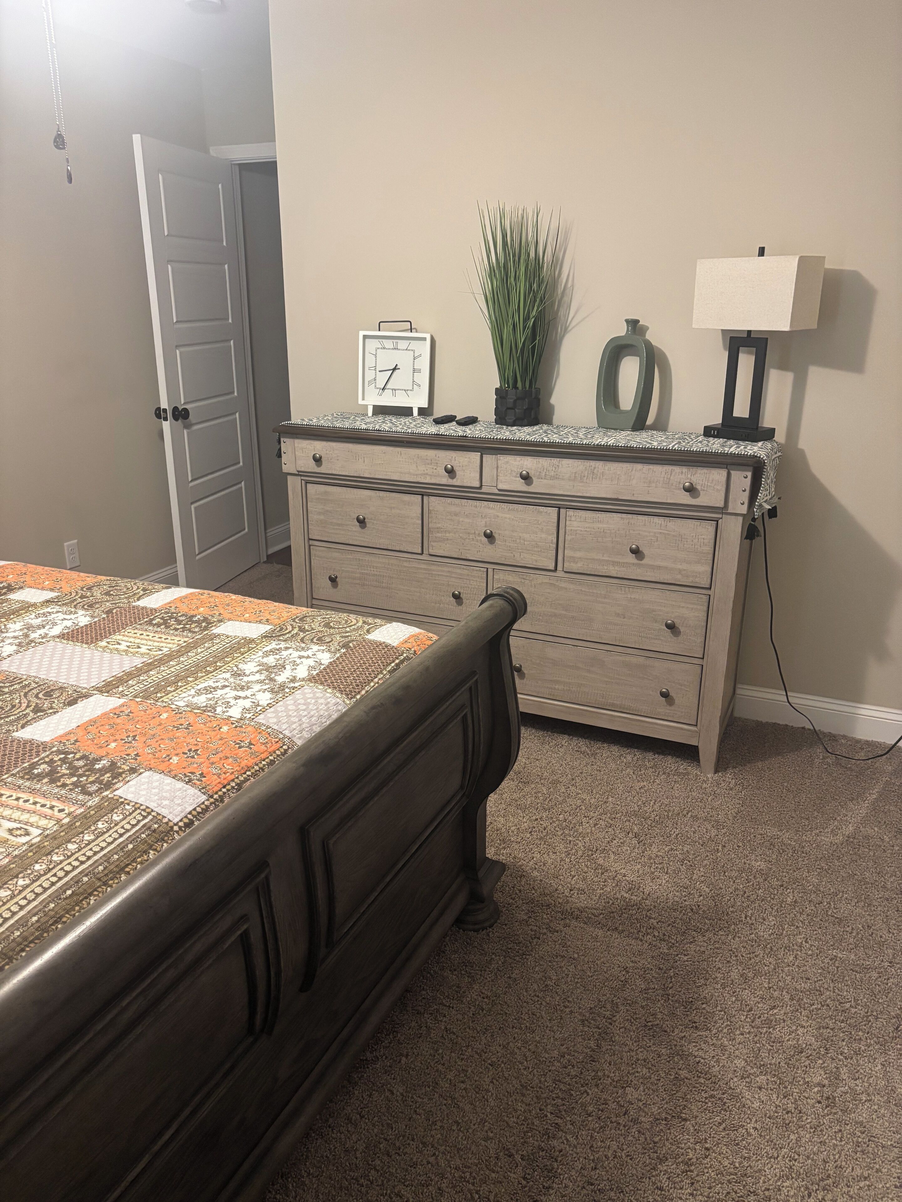 A bedroom with a bed, dresser, clock, plant, and lamp.