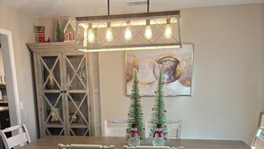 A dining area with a wooden table, two chairs, a chandelier, a decorative cabinet, and two Christmas trees.