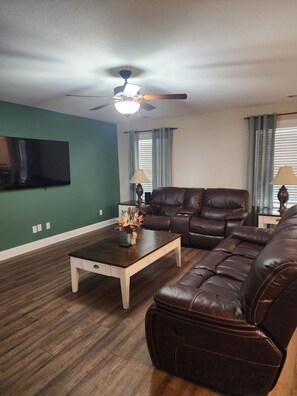 A living room with a ceiling fan, a flat-screen TV, a brown leather sofa, a wooden coffee table, and a lamp.