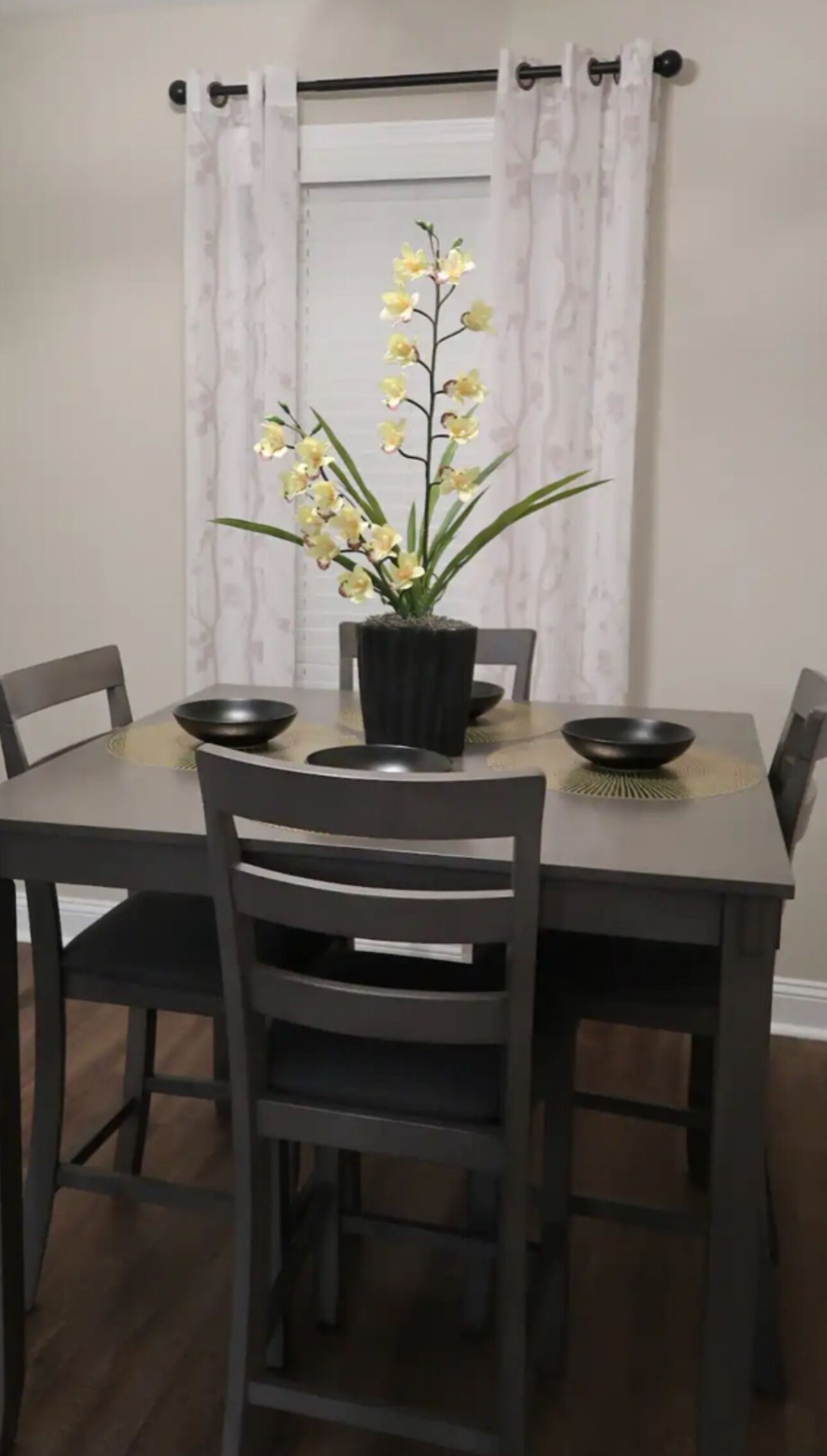 Een eettafel met vier stoelen, een zwarte vaas met gele bloemen, en een raam met witte gordijnen.
