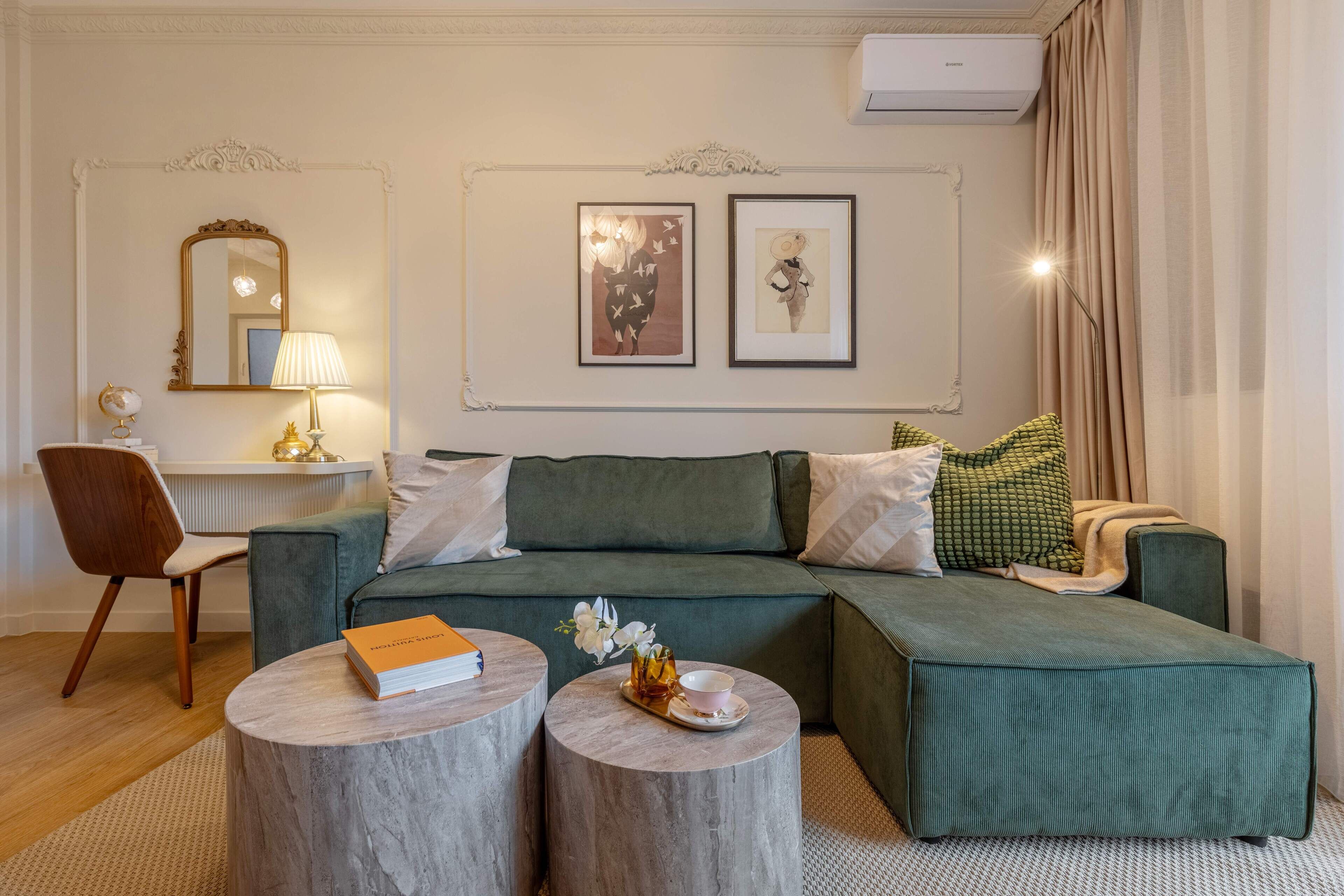 A modern living room with a green sectional sofa, two round wooden side tables, a mirror, and two framed artworks on the wall.