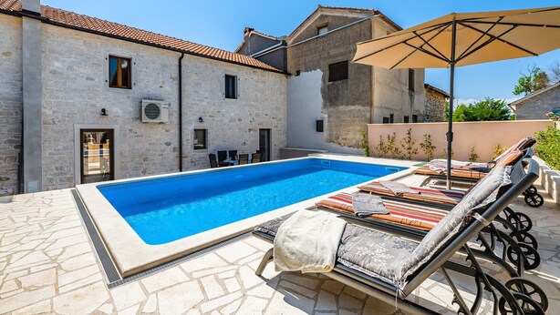 A swimming pool with lounge chairs and an umbrella, stone walls, and a clear blue sky.