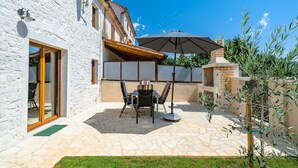 A stone patio with a table and chairs, a brick oven, and a white building with a wooden door.