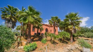 A rustic building with a red facade, surrounded by palm trees and cacti, set in a dry, rocky landscape.