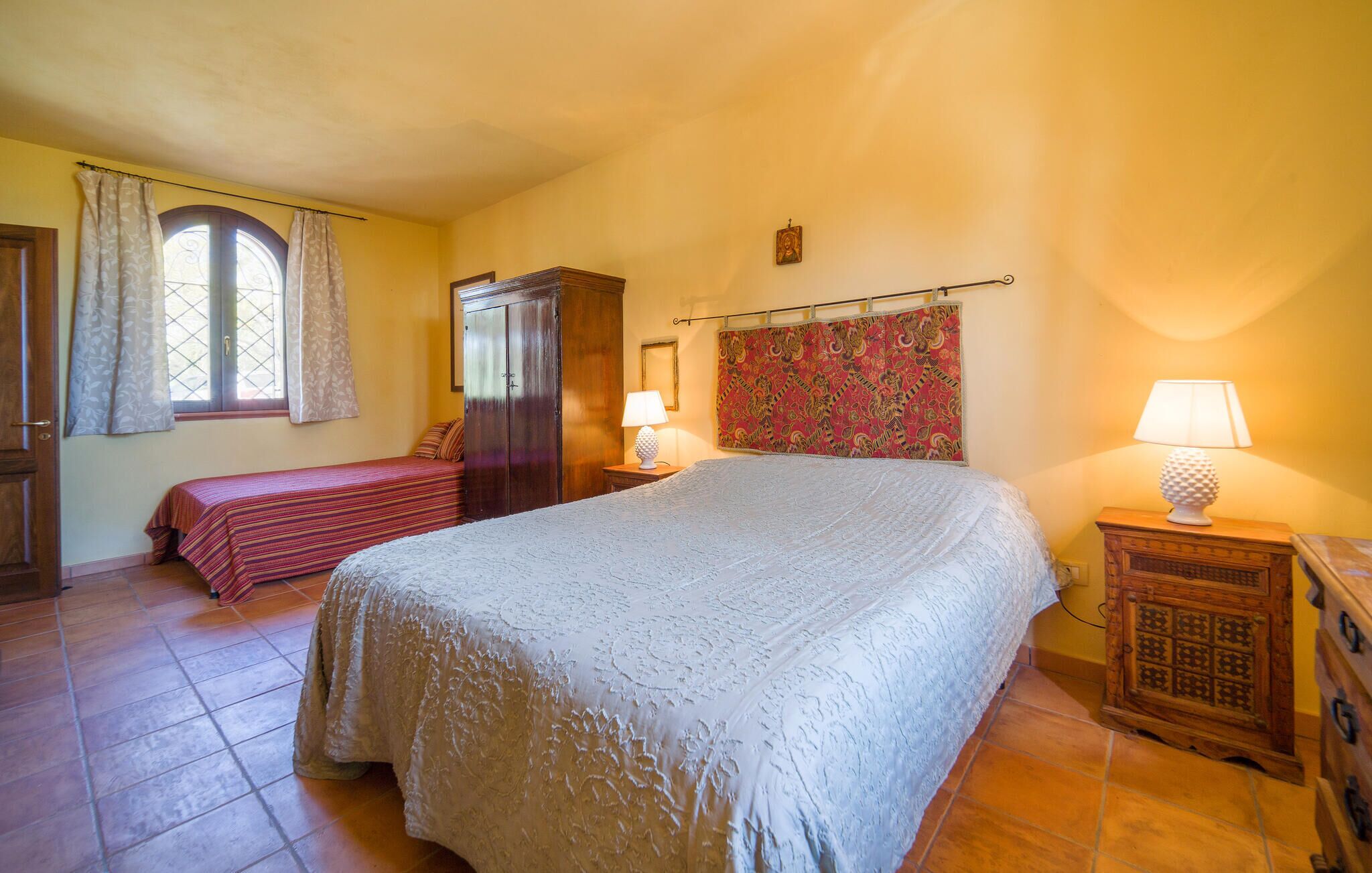 Une chambre avec un grand lit, un lit plus petit, une armoire, une fenêtre avec des rideaux et une tapisserie à motifs sur le mur.