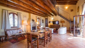 A spacious living room with a wooden ceiling, a fireplace, a dining area with a table set for four, and a staircase leading to an upper level.