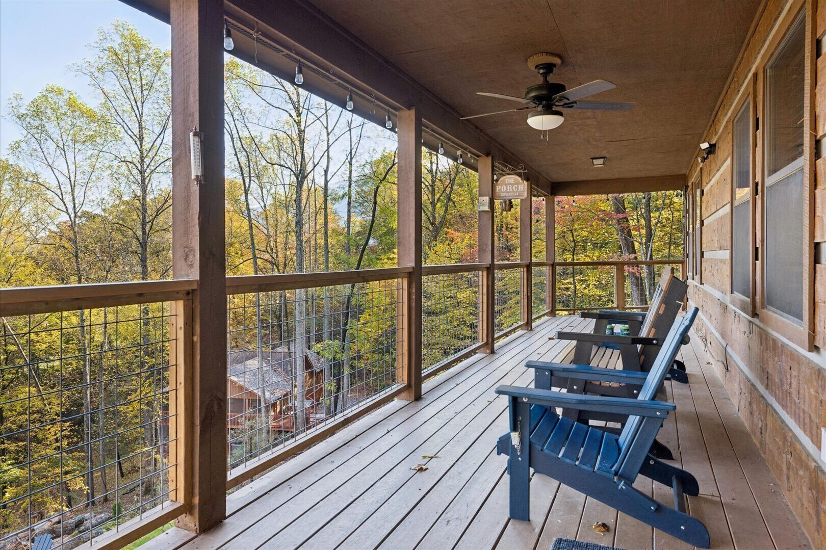 A covered porch with wooden flooring, blue Adirondack chairs, and a view of autumn trees.