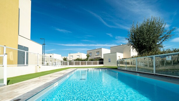 A swimming pool with clear blue water, surrounded by modern buildings and a tree. - Sweet home duplex 6 pers (Sérignan)