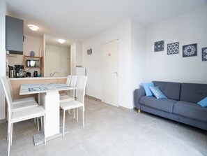 A modern living room with a grey sofa, white chairs, a dining table with a magazine, and a kitchen area with a microwave and coffee machine.