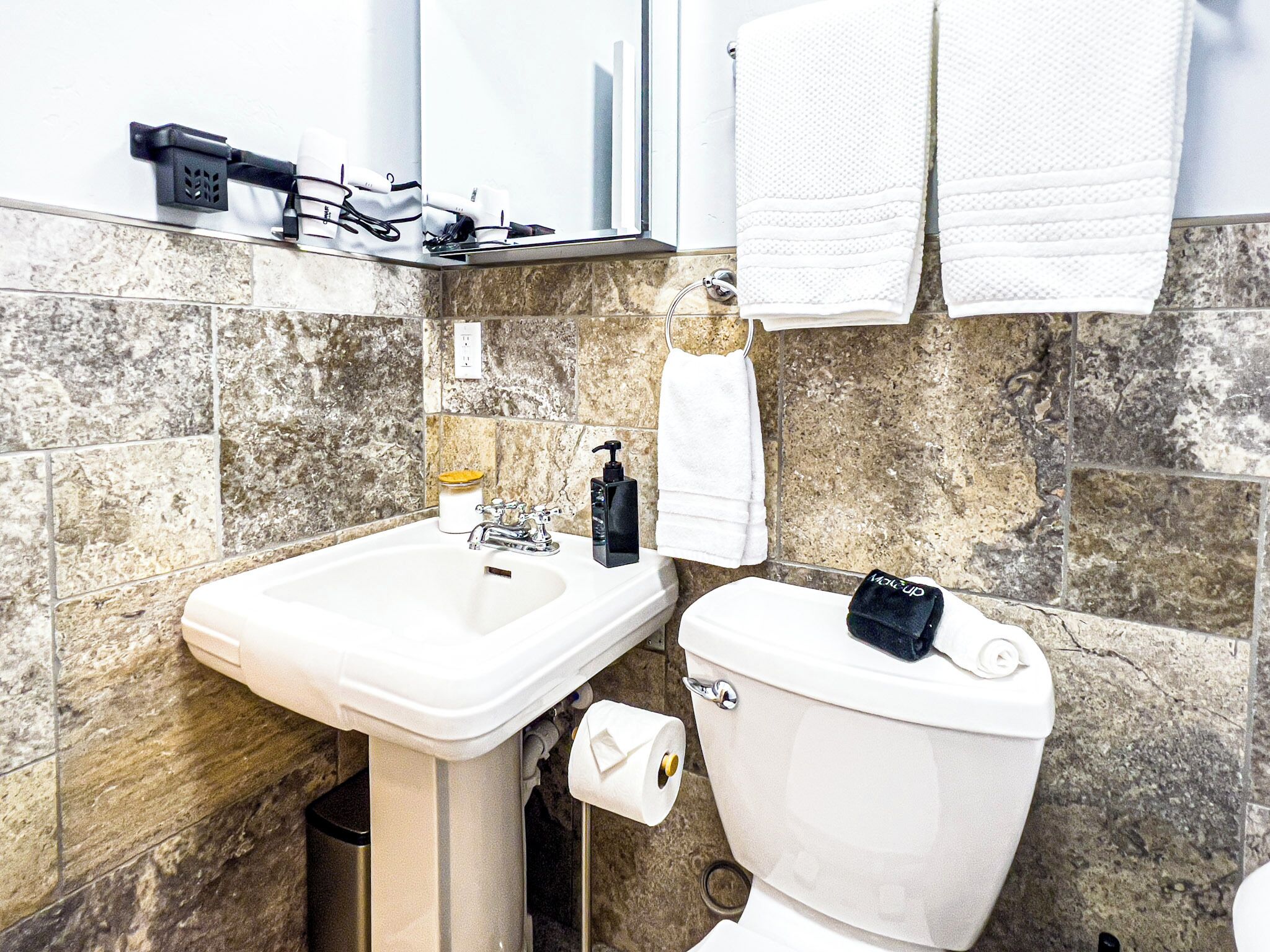 Een badkamer met een wastafel, toilet en spiegel. Handdoeken hangen aan de muur en er is een zeepdispenser op de wastafel.