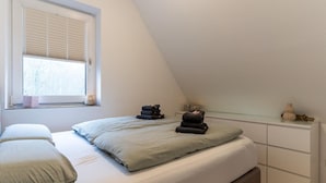 A bedroom with a bed, pillows, a bedside table, and a window with blinds. - Ferienwohnung in St. Peter-ording (St. Peter-Ording)