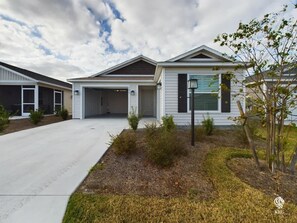 A modern two-story house with a garage, a covered driveway, and a landscaped front yard with small shrubs and a young tree. - Perfectly Placed in Moultrie Creek (Sumter County)