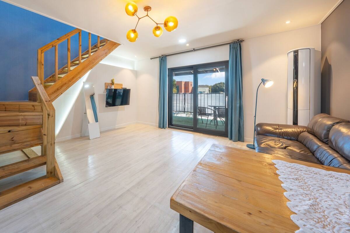 A modern living room with a wooden staircase, a flat-screen TV, a leather sofa, and a large window with a view of the outside.