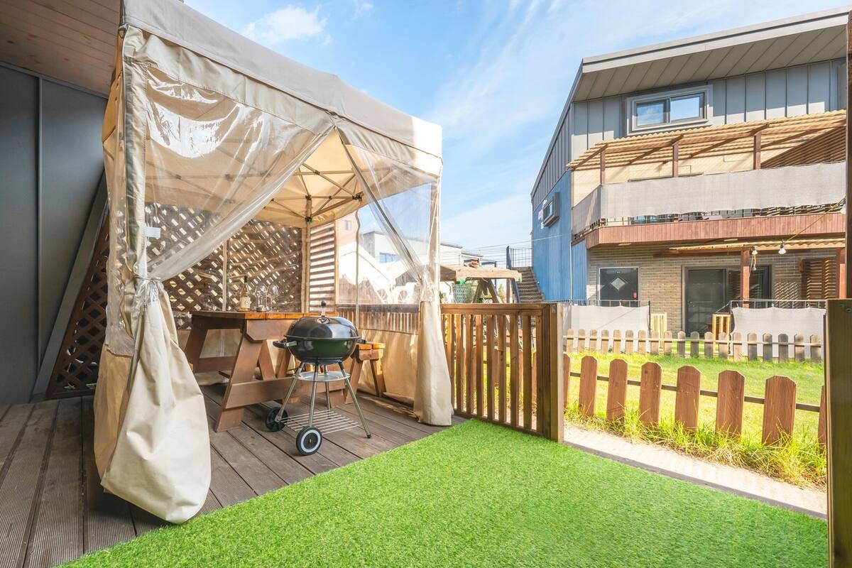 A covered patio area with a grill and a table, surrounded by a wooden deck and a small lawn.