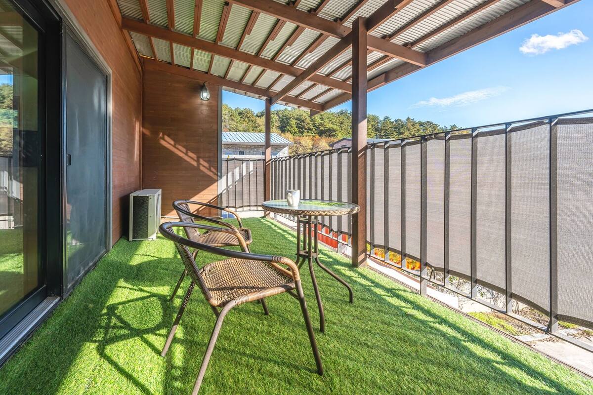 A balcony with artificial grass, a table, two wicker chairs, and a metal fence.