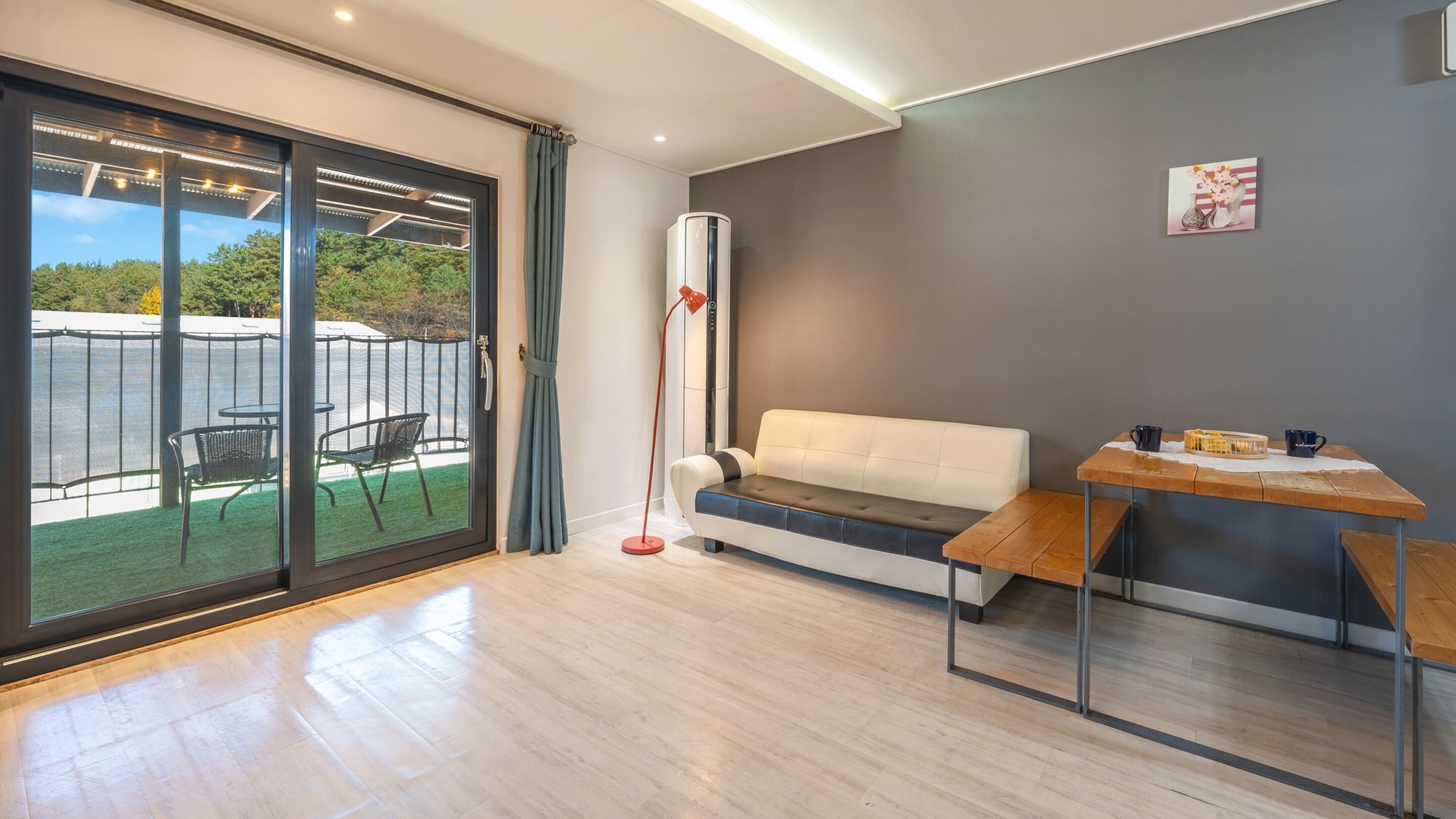 Una sala de estar moderna con un ventanal corredera que da a un balcón, un sofá blanco, una mesa de madera y una lámpara de pie.