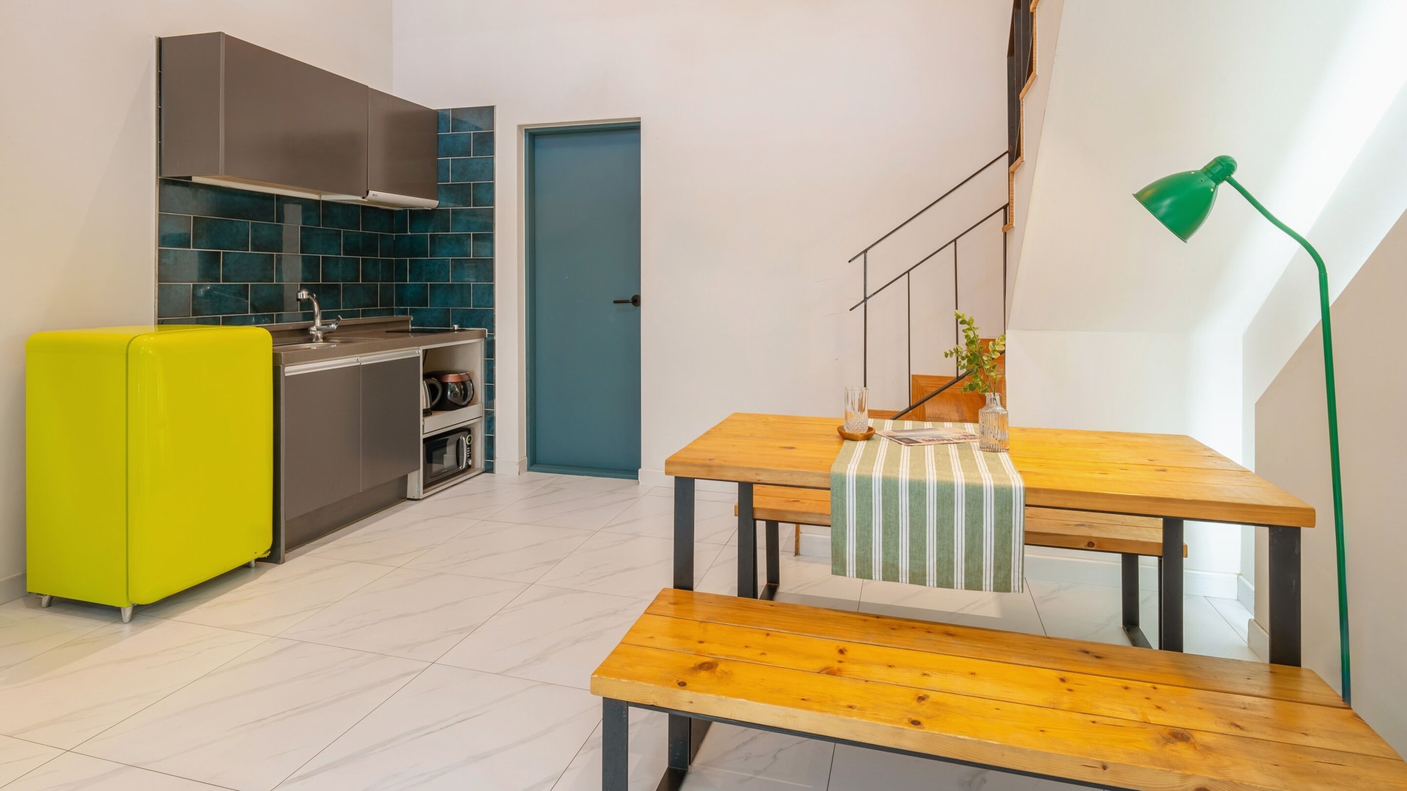 Une cuisine moderne avec un réfrigérateur jaune vif, une table à manger en bois recouverte d’une nappe à rayures et un lampadaire vert.