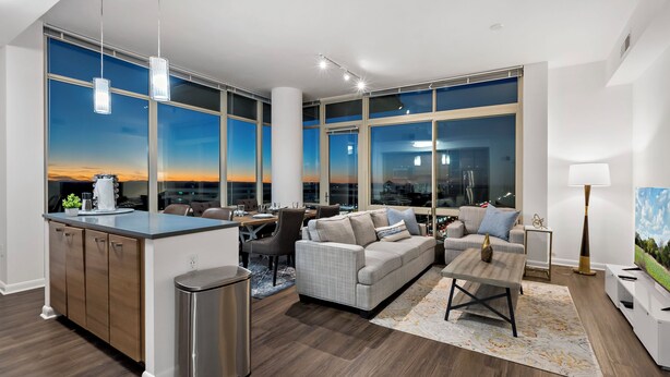 A modern living room with a dining area, large windows with city views, a kitchen with a bar area, and a cozy seating arrangement. - Tysons 1BR + Queen Den | Rooftop Pool & Balcony (Tysons)