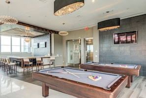 Une table de billard avec queues et billes, un coin bar avec des tabourets et une grande fenêtre laissant entrer la lumière naturelle.