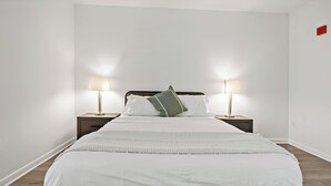 En pænt redt seng med hvidt sengetøj og en grøn pude, omgivet af to sengeborde med lamper, i et rum med hvide vægge og trægulv.