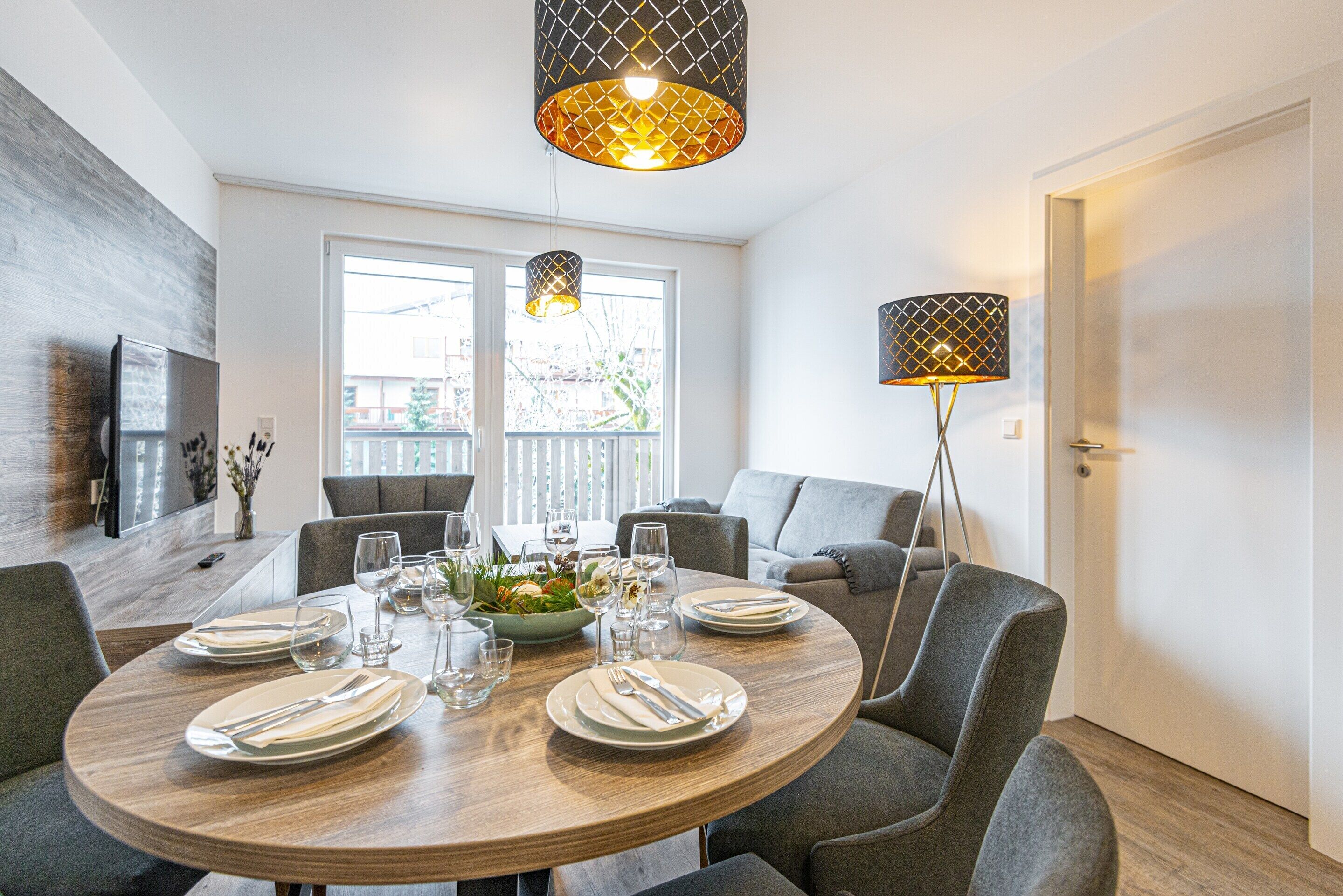 A dining area with a round table set for four, surrounded by grey upholstered chairs. There are two pendant lights above the table, and a floor lamp to the right. The room has a modern design with a wooden accent wall and a large window providing natural light.