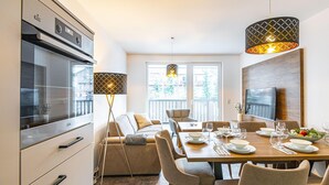A modern kitchen with a built-in oven, a dining area with a wooden table set for four, and a large window with a view of trees. - Ideal Apartment in Salzburg Without Deposit (Bad Hofgastein)