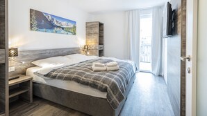 A modern hotel room with a large bed, a painting of a mountain landscape, a TV mounted on the wall, and a balcony with curtains. - Ideal Apartment in Salzburg Without Deposit (Bad Hofgastein)