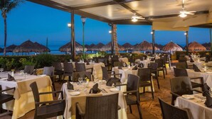 Zona pranzo all'aperto con tavoli e sedie, ventilatori a soffitto e vista sulla spiaggia al tramonto.