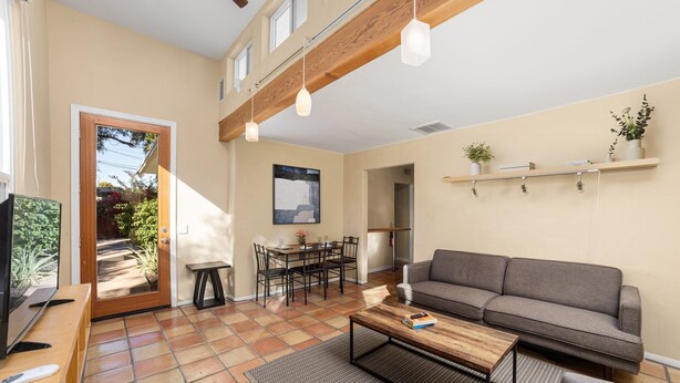 Ein Wohnzimmer mit Sofa, Couchtisch und Fernseher. Es gibt einen Essbereich mit Tisch und Stühlen. Der Raum hat eine Holzbalkendecke und eine Tür, die nach draußen führt.