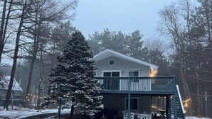 A two-story house with a balcony, surrounded by snow-covered trees and a small deck with outdoor furniture.