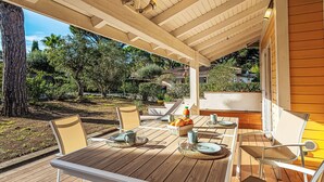A covered outdoor patio with wooden tables and chairs, a small table with fruit and a pitcher, and a view of a garden and buildings in the background. - Parc Oasis by Interhome (Gassin)