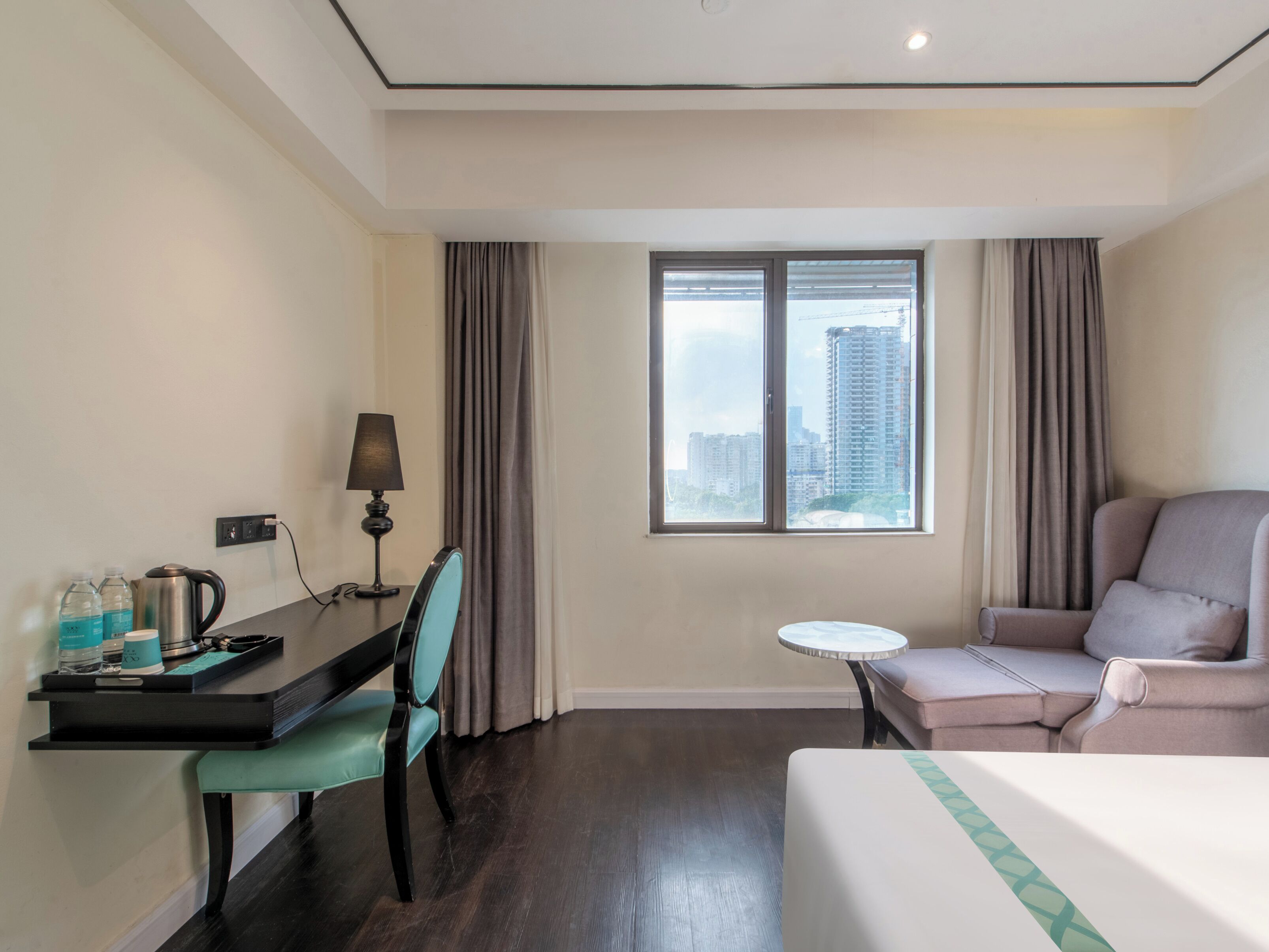A hotel room with a bed, a desk with a chair, a small table, and a view of buildings through the window.