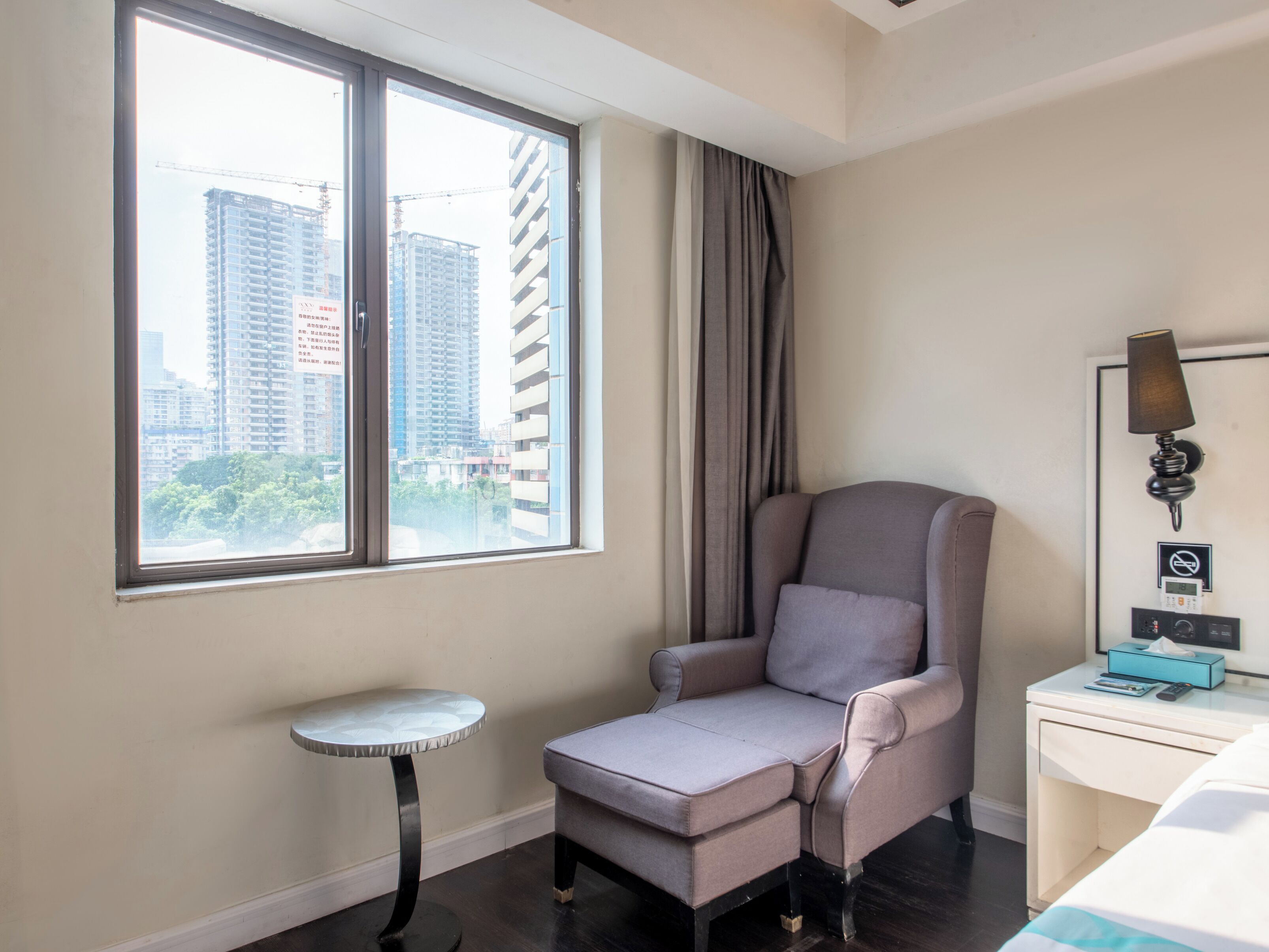 Habitación de hotel con un ventanal grande, un sillón gris, una mesita redonda pequeña, una mesita de noche con lámpara y una cama.
