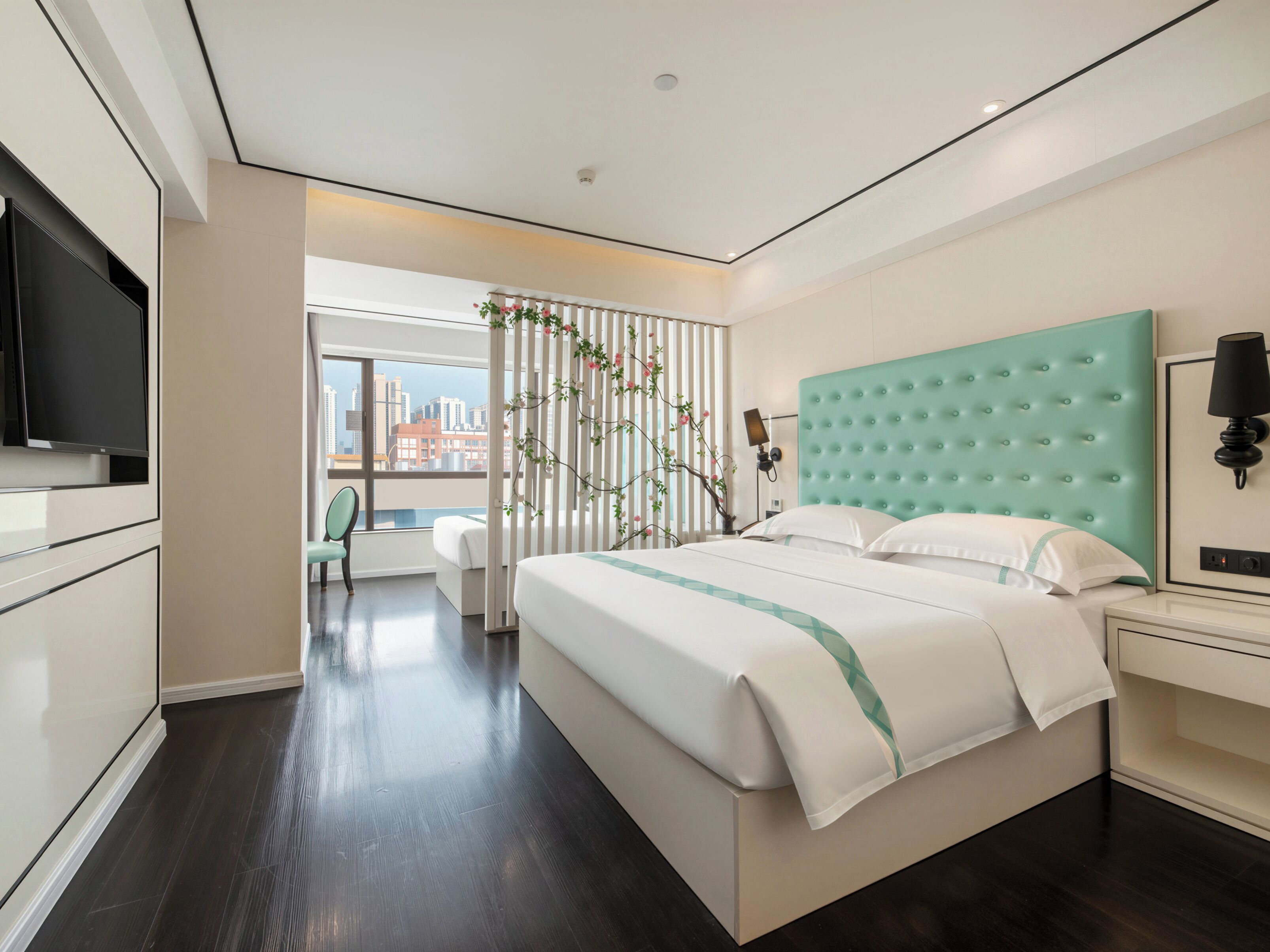 A modern hotel room with a large bed, a television, and a view of the cityscape.