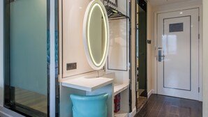 A bathroom with a white sink, a blue chair, a mirror with lights, and a door in the background.