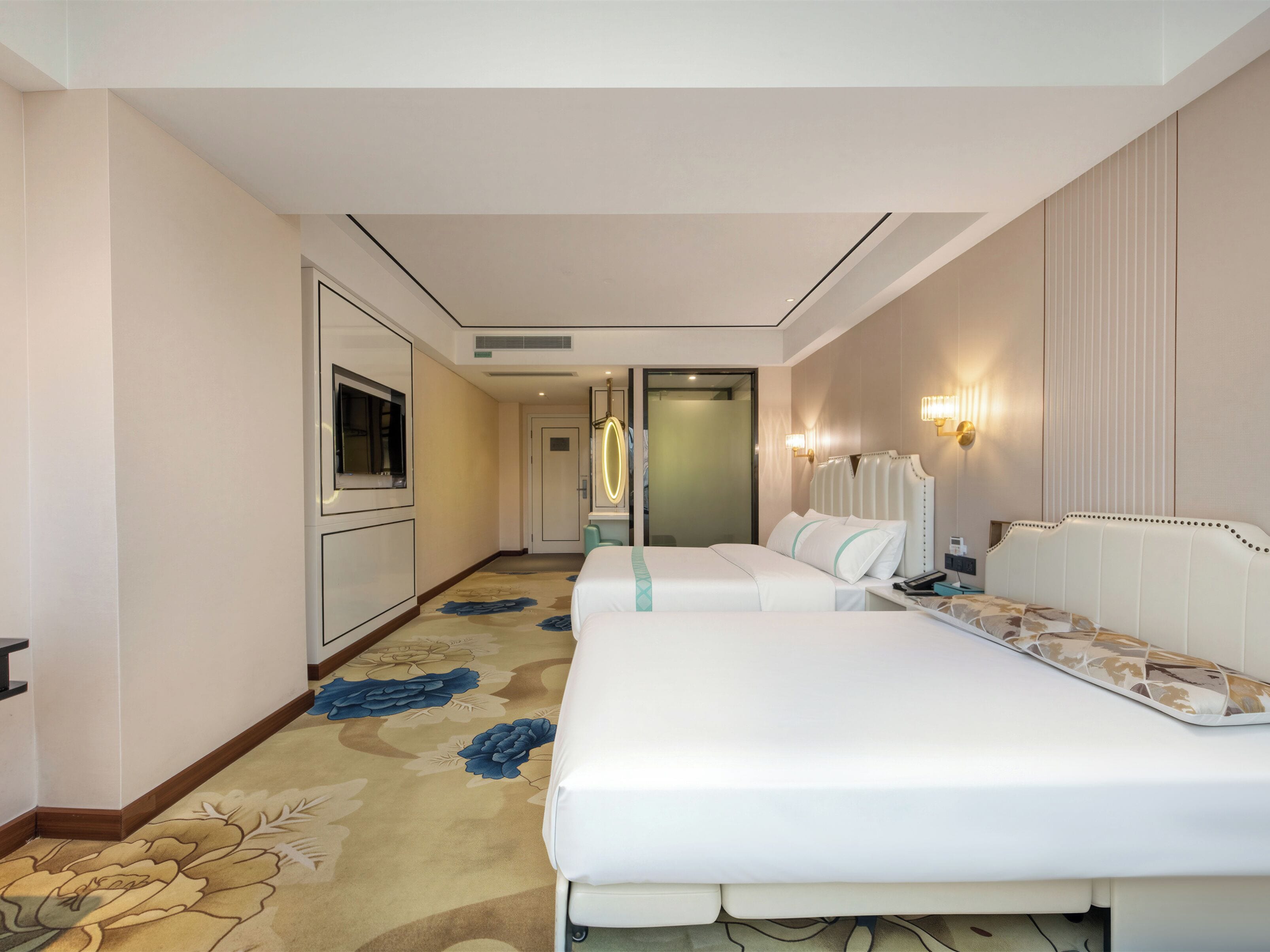 A hotel room with two beds, a large mirror, a television, and a carpet with a floral pattern.