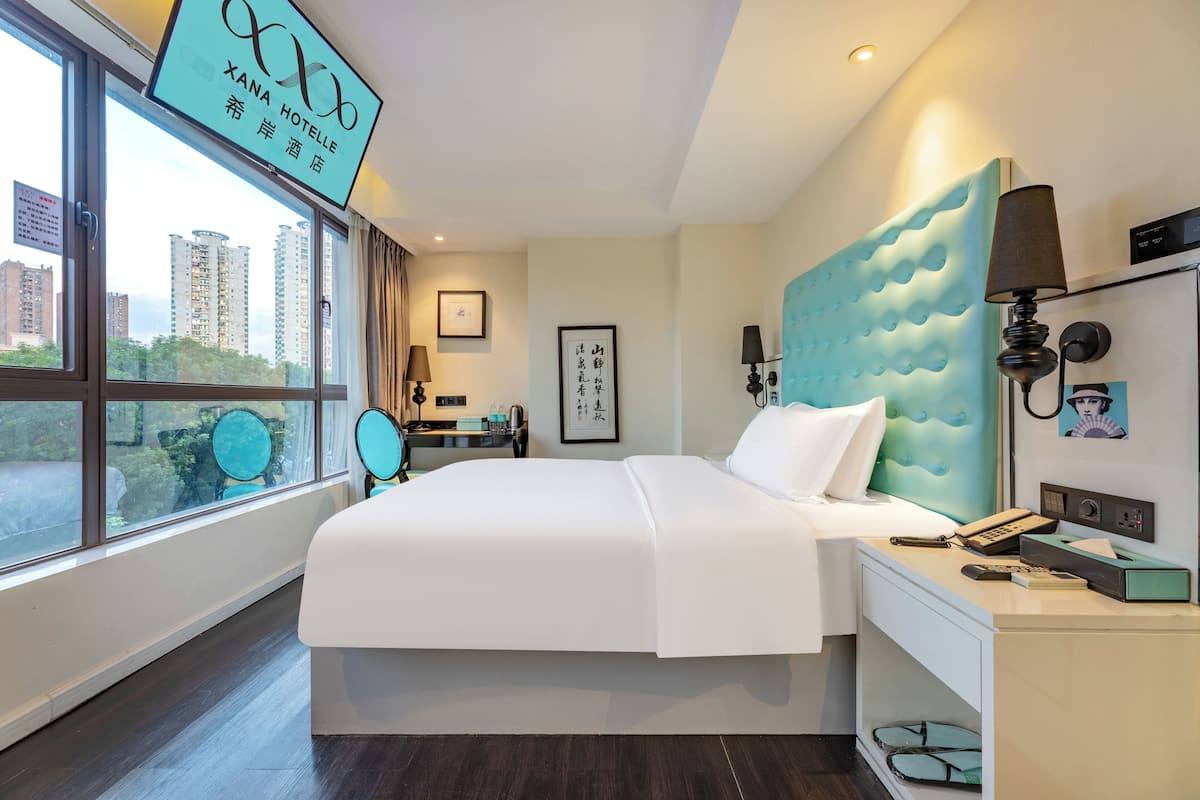 Een hotelkamer met een groot bed, een nachtkastje met een lamp, een raam met uitzicht op hoge gebouwen en een bord boven het raam.